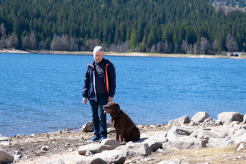 A senior man with dog is staying on the bank near the blue lake