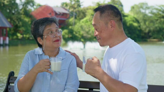 A man and a woman are sitting on a bench by a lake. The man is holding a cup of milk and the woman is holding a cup of coffee. They are both smiling and seem to be enjoying each other's company