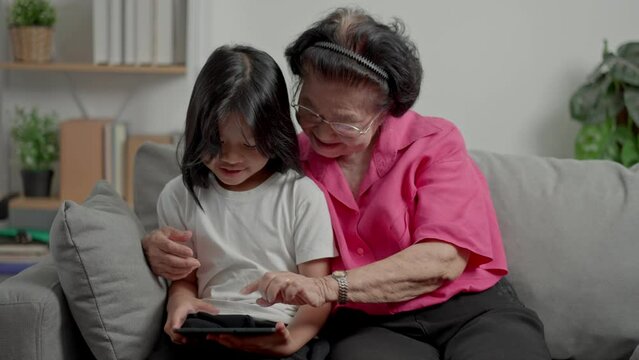 Smiling overjoyed mature grandmother with little granddaughter using tablet on cozy couch. Happy mature granny and small grandchild play education videos tablet. Education online, mature joy concept.