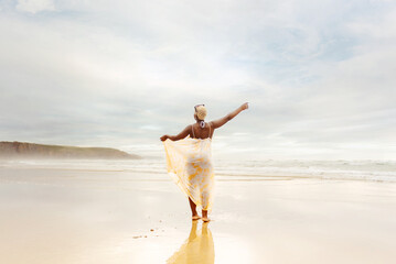 curvy black woman poses on the beach stretching her arms and gazing at the sea in a slim summer dress