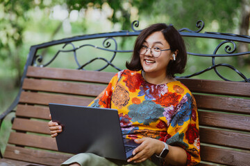 Female professional enjoys remote work on a calm sunny day in a lush green park An Asian businesswoman smiling while successfully completing tasks on laptop on bench outdoors