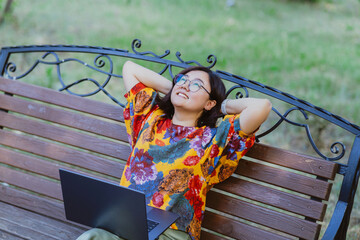 Asian freelancer with laptop enjoying relaxing on bench in park in summer Freelance lifestyle portrayed by a woman engaging with technology in a peaceful park setting