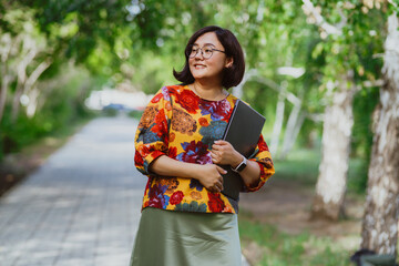 A smiling Asian woman with glasses holding a laptop in a sunny park. Happy entrepreneur enjoying remote work amidst green trees outdoors