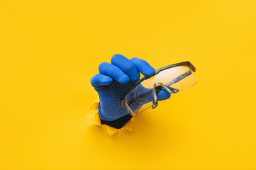 The right male hand in a blue fabric work glove holds safety glasses. Torn hole in yellow paper. Good job, eye protection and safety concept. Copy space.