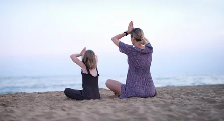 Schilderijen op glas Mother and daughter do yoga meditate in lotus position on beach. Family yoga class by sea concept © H_Ko