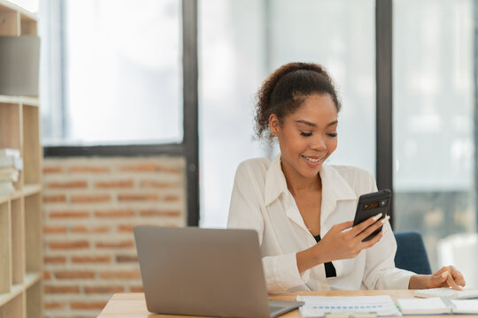 Young african american businesswoman smiling while checking messages on smartphone or browsing social media, working at desk with laptop and documents in modern office - Powered by Adobe
