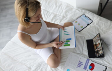 Woman in headphones holds charts sits on bed and holds online conference. Remote work and freelancer