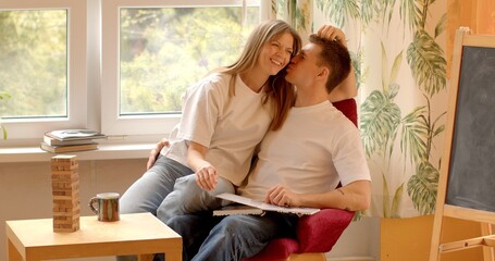 Couple kisses while sitting in a chair at home. Young couple posing, smiling, embrace symbolizes...
