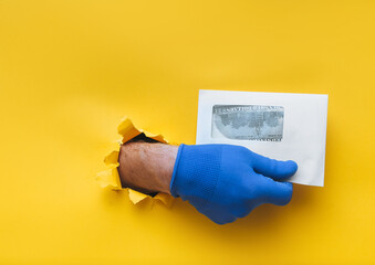The male hand in a blue fabric work glove holds an envelope with dollar bills (money). Torn hole in yellow paper. Concept of guest worker, handyman, gray illegal salary for work. Copy space.