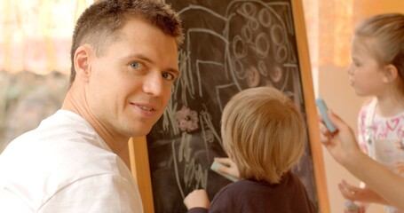Man looks into the camera against the backdrop of children drawing in a preschool. Early childhood...