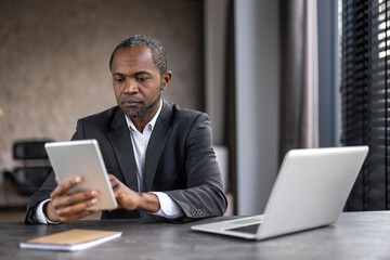 A man in a suit is sitting at a desk with a tablet and a laptop. He is focused on the tablet, possibly checking emails or browsing the internet. Concept of productivity and concentration