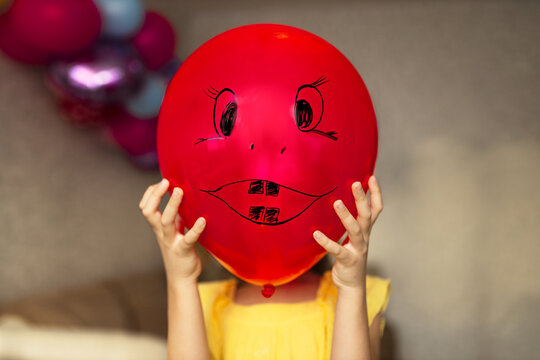 Girl in yellow dress covers her face of red balloon with drawn smiley face in holiday at home.
