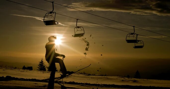 Silhouette of a female skier against sunset sky beneath a row of empty ski lift chairs. Girl throwing show in the air with her ski. 120fps cinema camera.