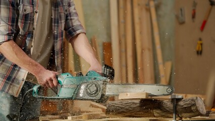 Male woodworker working in garage. Man professional carpenter working with wooden materials in workshop sawing wood using electric chainsaw.