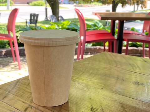 Biodegradable, compostable coffee cup on a city bistro table. Cardboard goblet for a hot beverage on a wooden table in a garden. Restaurant terrace, red chairs and greenery in summer.