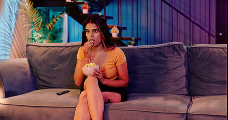 Obraz premium Indian Asian Hindu gen z young woman sitting sofa couch watch horror scary movie indoor home scared adult afraid girl hold bowl eat snacks look fearful series film on TV enjoy fun joy late night house