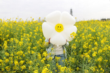 Young woman holds daisy balloon with smiling face in front of own face in the middle of a rapeseed (Brassica napus) field full of bright colourful yellow flowers. Spring, summer, happy, joy concept