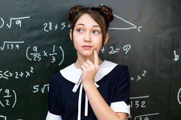 A teenage schoolgirl is thinking while standing at the blackboard, a 10-12 year old girl is solving a math problem.