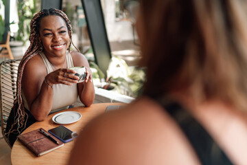 young African-American woman shares joyful moment with companion at cozy café. She's holding...