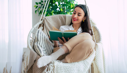 Happy lovely relaxed trendy woman with long healthy hair reads a book in stylish clothes in the modern living room while sitting on hanging chair. - 781407804