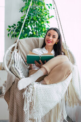 Happy lovely relaxed trendy woman with long healthy hair reads a book in stylish clothes in the modern living room while sitting on hanging chair. - 781407819