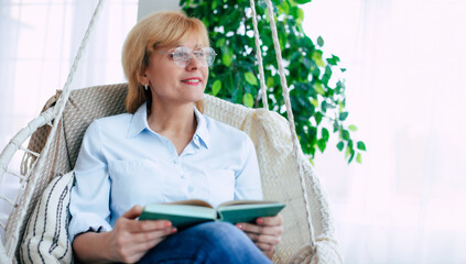 Close up photo of relaxed mature or middle-aged woman retired in eyeglasses reading a book in the modern living room while sitting on hanging chair and thoughtful about something.