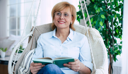 Close up photo of relaxed mature or middle-aged woman retired in eyeglasses reading a book in the modern living room while sitting on hanging chair and thoughtful about something. - 781407860