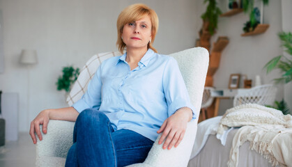 Home interior image of sad thoughtful mature woman looking on camera while sitting at home on the chair - 781408022