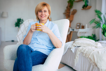 Happy lovely mid-aged woman resting with a smile while sitting with a cup of tea in her hands on the chair. Cheerful mature female enjoying a serene retirement at home. - 781408078
