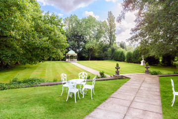 Deserted manicured garden with a gazebo at the end of stone path through a lawn surrounded by trees...