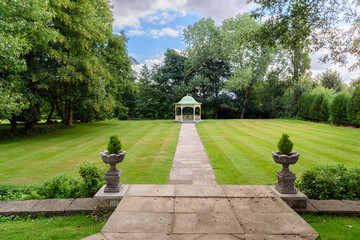 Deserted gazebo at the far end of a stone path through a lawn surrounded by trees in a park on partly cloudy summer day