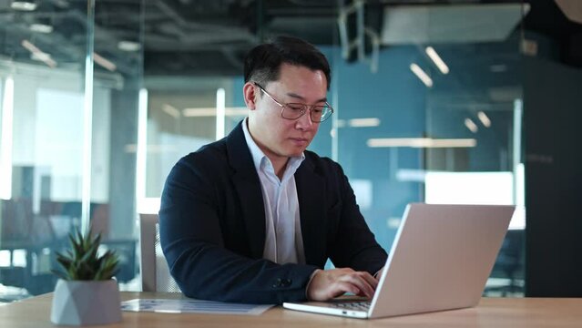 Attentive male accountant wearing glasses quickly typing text on laptop while creating financial report. Concentrated man in suit writing letter on computer while sitting at desktop in office.
