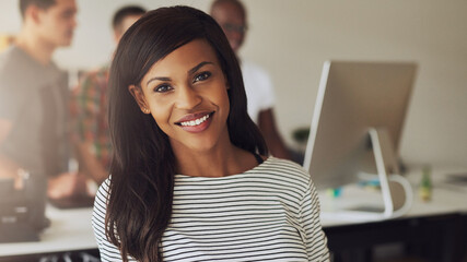 A young african american confident female entrepreneur sits in an office and looks focused into the camera while smiling. Her colleagues stand behind her and look at a computer screen