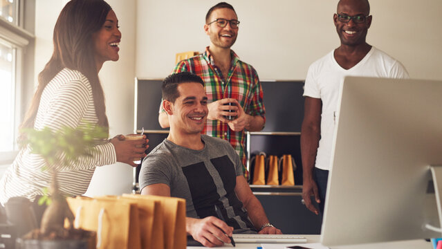 Man showing something on his computer to his three diverse colleagues. They laughing at each other and are happy about what they see on the screen