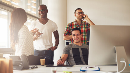 Four young diverse entrepreneurs stand together in an office community. A man looks into the camera smiling while the others talk together and talk on mobile phones - obrazy, fototapety, plakaty