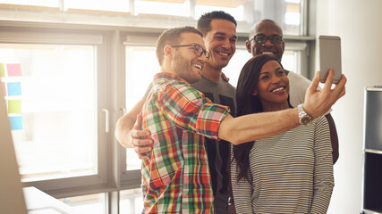 Four happy young diverse entrepreneurs stand together in an office community. They take a selfie with a mobile while they are all smiling and laughing.