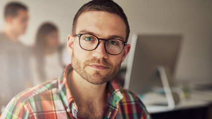 A young entrepreneur man sits in an office and looks focused into the camera, while his colleagues...