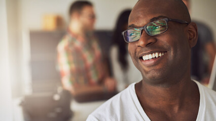 Happy black man with black glasses, smiling while his diverse colleagues stand in the background and look at a computer screen.