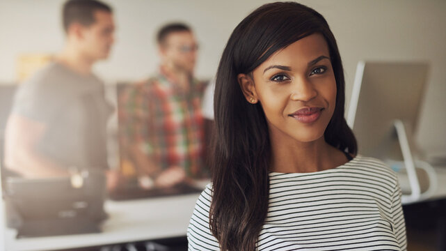 A young black female entrepreneur sits in an office and looks focused into the camera while smiling. Her diverse colleagues stand behind her and look at a computer screen