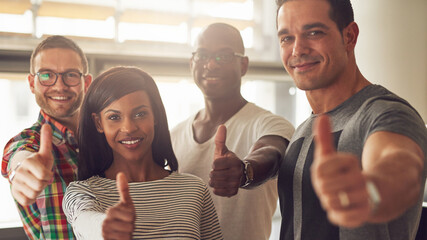 A group of diverse casually dressed business people stand together and look smiling directly into the camera while giving thumbs up. 