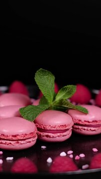 Pink sugar granules falling on rotating fresh macaron cookies. Traditional french desserts from almond dough. Pink macaron cakes, raspberry fruits and green mint leaves against black background.
