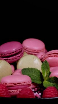 Fresh macaron cookies, raspberry fruits, pink sugar granules and green mint leaves on a plate. Traditional french desserts from almond dough. Beautiful macaron cakes rotating against black background.
