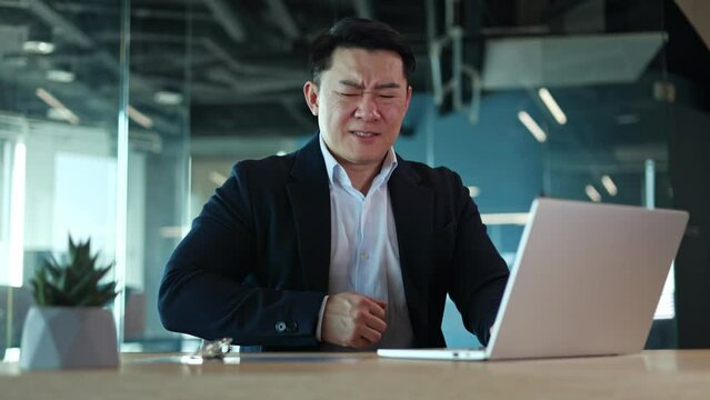 Concentrated asian male typing on laptop in office and suffering from sudden stomach ache. Company director in formal wear holding belly and experiencing acute attack of pain in abdomen during work.