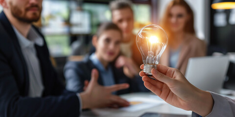 A hand holding a light bulb with business people in an office brainstorming meeting. A creative team or group with Innovative business idea. - obrazy, fototapety, plakaty