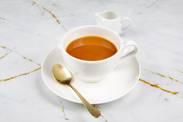 A cup of coffee with a small creamer and a golden spoon on a marble table
