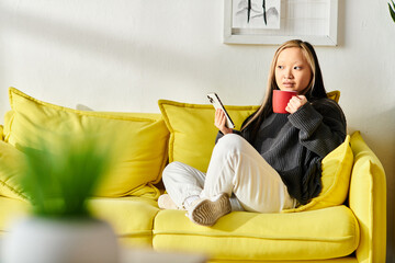 A young woman of Asian descent sits on a yellow couch, holding a cup of coffee in her hands while...