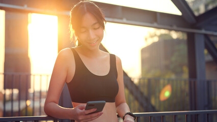 Fitness happy asian woman using smartphone and resting after running on city in the morning, sunset