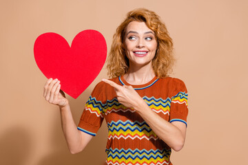 Photo portrait of lovely young lady hold red heart look point postcard dressed stylish striped garment isolated on beige color background