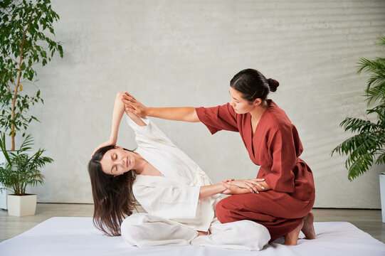 Masseuse making thai yoga massage. Woman therapist doing traditional massage treatment, stretching female patient's body. Client sitting on mat, enjoying hand massage in calm atmosphere.