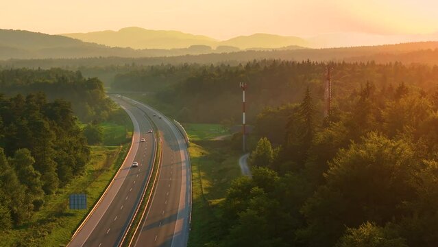 AERIAL, LENS FLARE: Evening sunlight spills over scenic landscape and winding highway. Cars and trucks drive through a beautiful countryside where gentle mists create an incredible painterly scenery.